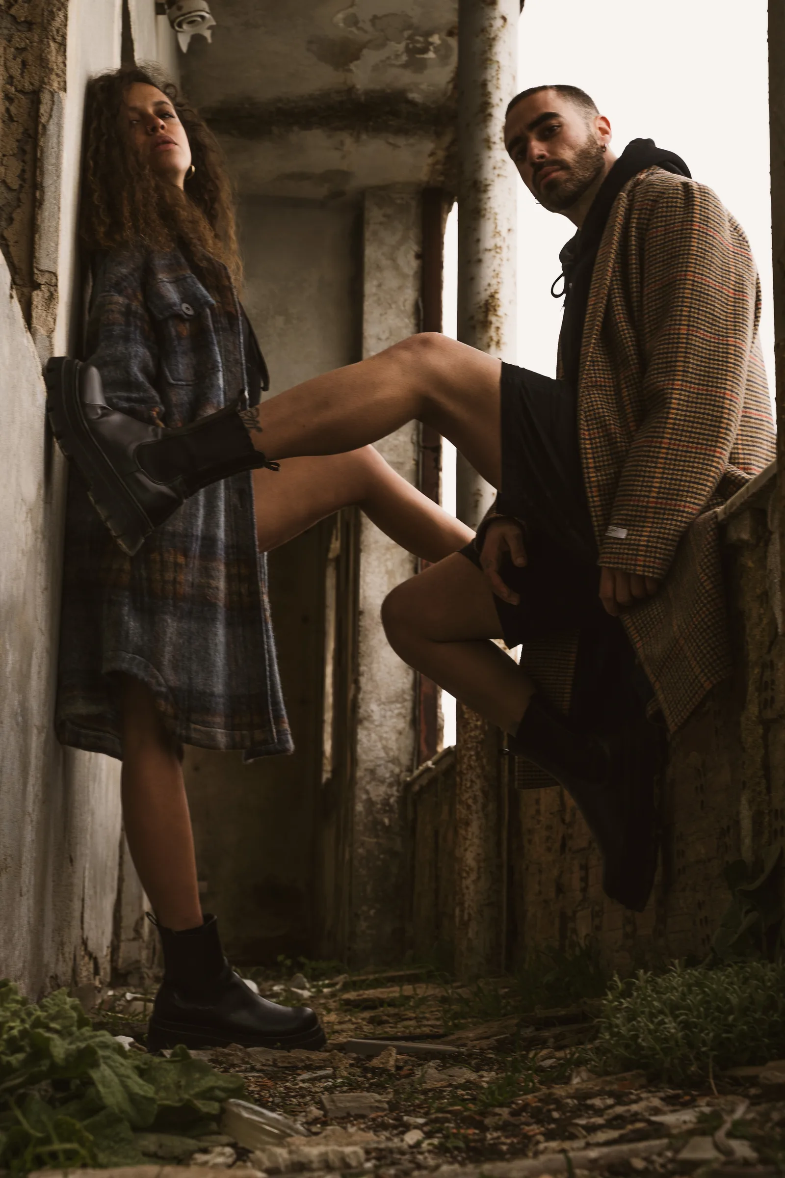 Two models in oversized coats leaning on a weathered wall