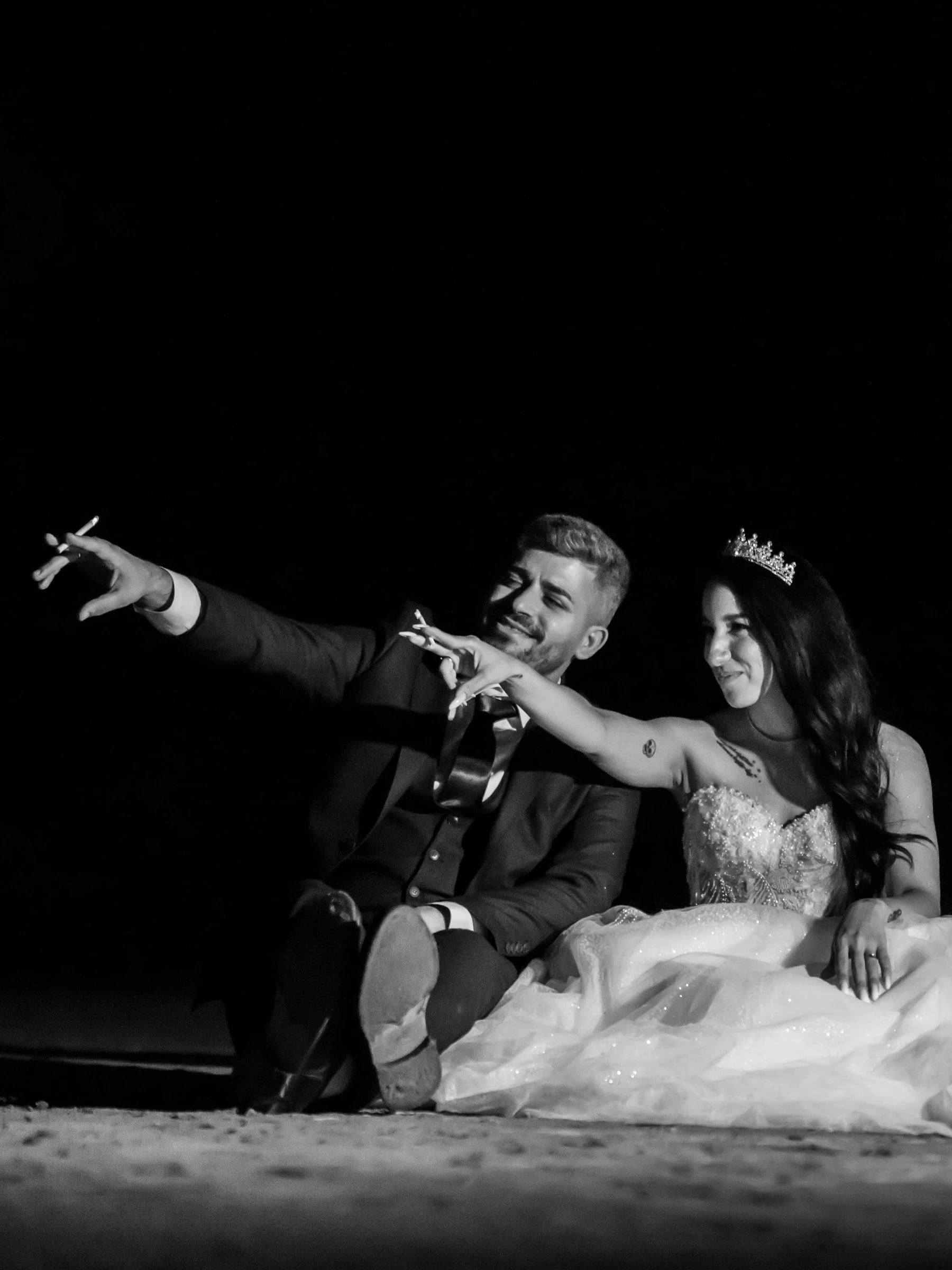 Bride and groom sitting on the ground at night, laughing and pointing into the distance